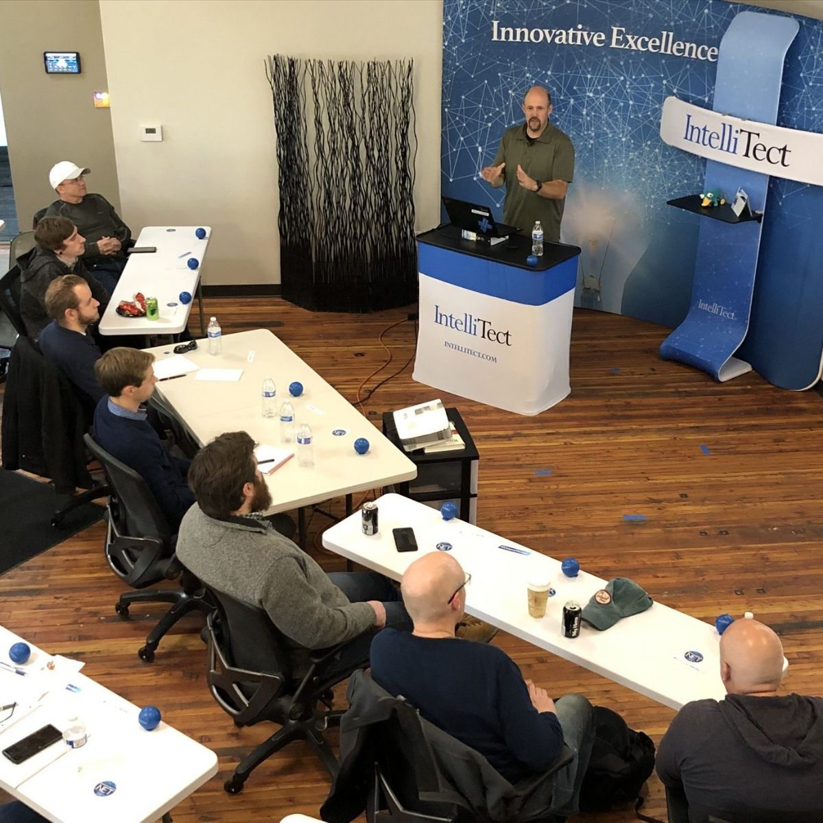 Grant Erickson, IntelliTect CTO, stands behind a white and blue podium presenting to a room of developers seated at white tables