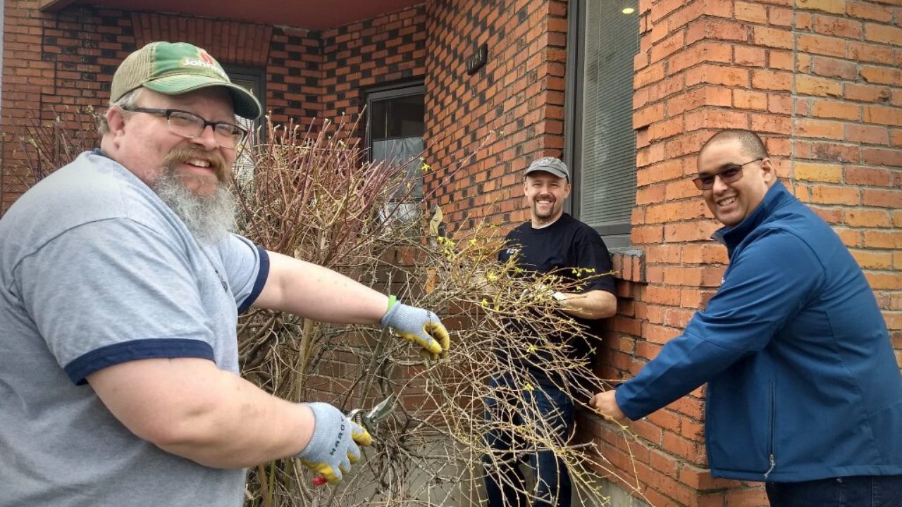 Three smiling men work to trim bushes and clean the yard in front of a red brick building